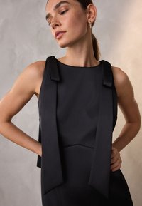 Musta hihaton paita, jossa on solmukkeella koristellut olkapäät, siinä on sileä pinta ja istuva muotoilu, korostaen minimalistista estetiikkaa.