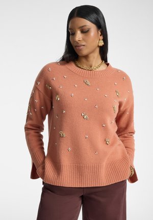 Frau trägt einen lachsrosa Pullover, verziert mit kleinen Perlen- und Goldapplikationen, kombiniert mit dunkelbrauner Hose und goldenem Schmuck.