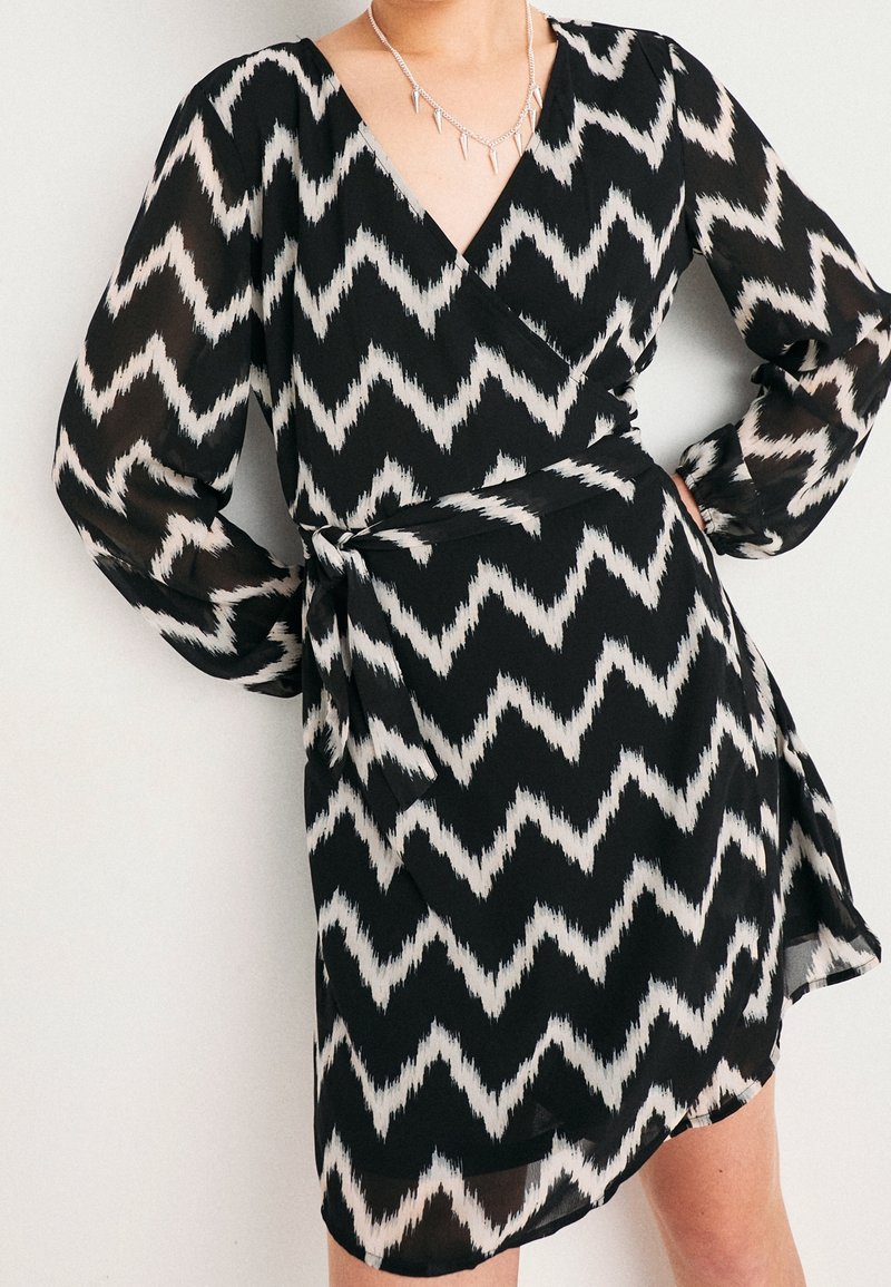 Femme portant une robe portefeuille noir et blanc à motif zigzag avec de longues manches transparentes et un collier pointu en argent.