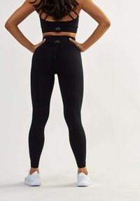 Leggings noirs taille haute avec une texture lisse, présentant un design ajusté et deux petits accents de logo à l'arrière, assortis à des baskets blanches.
