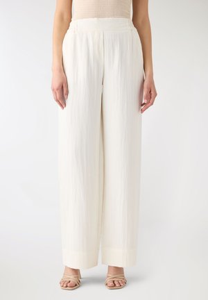 Donna che indossa pantaloni bianchi a vita alta e gamba larga abbinati a sandali beige con cinturini, in piedi davanti a uno sfondo bianco semplice.