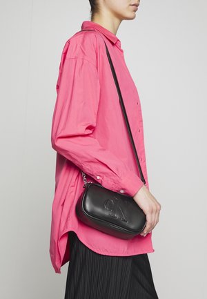 Vrouw in een loszittend roze shirt en een zwarte geplooide rok, die een kleine zwarte crossbody tas met CK-logo aan de voorkant vasthoudt.