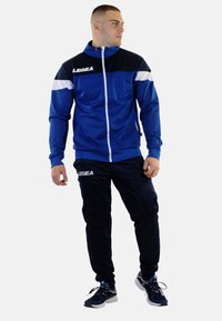 Blauwe sportieve trainingspak gemaakt van gladde stof; bevat een zipjack met witte accenten en logo, gecombineerd met zwarte broek en sneakers.