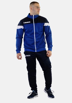 Chándal atlético azul hecho de una tela suave; presenta una chaqueta con cremallera y acentos blancos y un logo, combinada con pantalones negros y zapatillas.