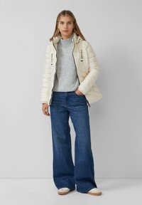 Witte gewatteerde pufferjack met een capuchon en zwarte branding, gedragen over een grijze gebreide trui en blauwe wijde jeans. Casual schoenen maken de outfit compleet.