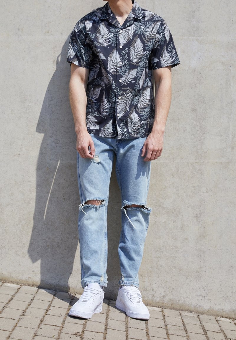 Zwart T-shirt met korte mouwen en een wit varenpatroon, gecombineerd met lichtblauwe ripped jeans en witte sneakers; staande op een verharde ondergrond.