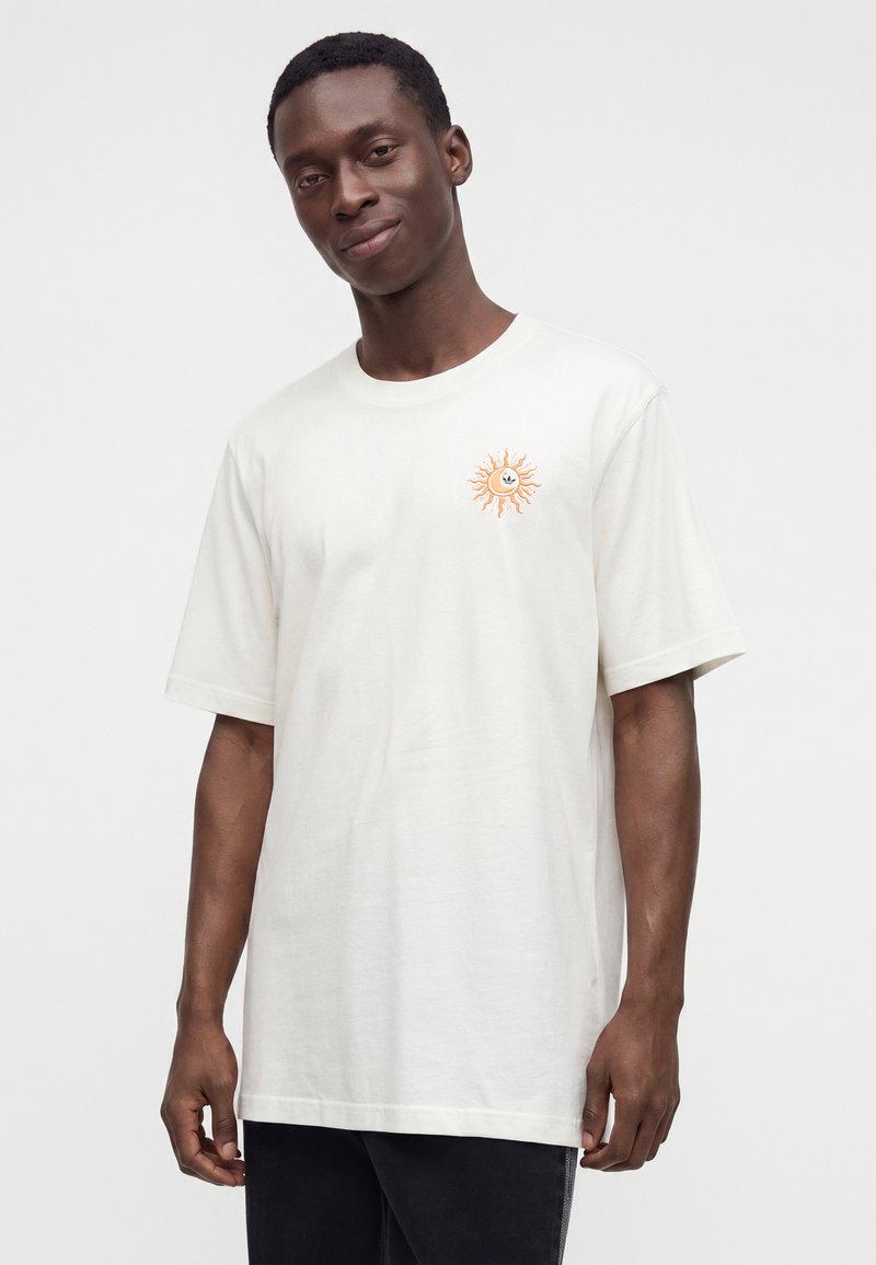 Uomo che indossa una t-shirt bianca a maniche corte con una piccola grafica di un sole arancione sul petto sinistro, su uno sfondo chiaro e uniforme.