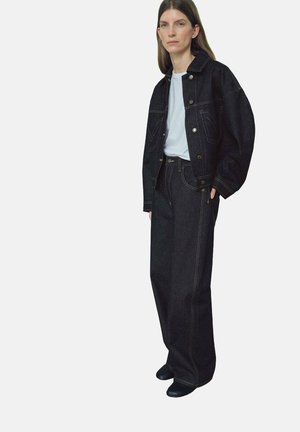 Chaqueta de mezclilla oscura con grandes bolsillos y costuras en contraste, combinada con jeans oscuros de pierna ancha y una camiseta blanca básica. Textura suave.