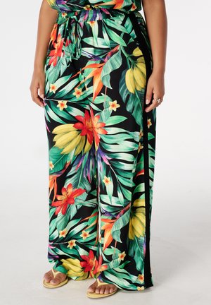 Wijde broek met bloemenprint met groene bladeren, rode en oranje bloemen en gele bananenclusters, gedragen met beige sandalen.
