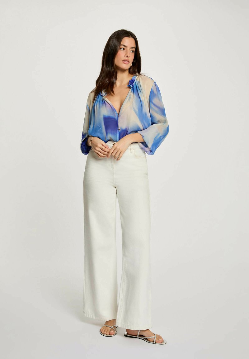 Donna in posa con una blusa ampia blu e beige, pantaloni bianchi a gamba larga e sandali argento con cinturini, su sfondo semplice.