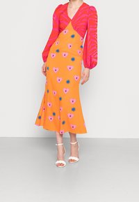 La robe présente un décolleté en V et de longues manches bouffantes, avec un corps orange vif orné de motifs de cœurs roses et d'étoiles bleues. Assortie à des talons blancs à lanières.