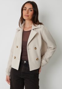 Beige dobbeltradet jakke lavet af blødt stof med slidskrave, prydet med store brune knapper og manchet detaljer, parret med en brun bluse.