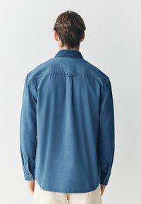 Blå skjorte med lange ærmer og knop-krave, der har en glat overflade og en afrundet bund. Bagerste udsigt viser skulder yoke og manchetter.