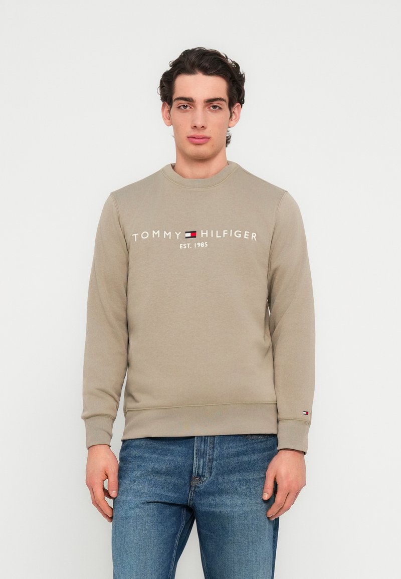 Μπεζ φούτερ με λαιμόκοψη crew, με λογότυπο "TOMMY HILFIGER" σε λευκό χρώμα, με κόκκινες και μπλε λεπτομέρειες, συνδυασμένο με μπλε τζιν.