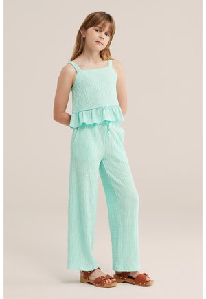 Młoda dziewczyna w teksturowanym, miętowo-zielonym topie bez rękawów z falbanką na dole i dopasowanych szerokich spodniach, brązowych sandałach, stoi z rękami za plecami.