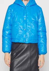 Person som bär en glänsande blå vadderad jacka med tryckknappar och en svart läderkjol, händerna avslappnade vid sidorna.