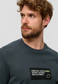 Czarnoszary bawełniany t-shirt z prostokątną grafiką z tekstem w czerni i bieli oraz żółtymi akcentami. Krótkie rękawy i okrągły dekolt.