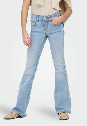 Person iført lyseblå vide jeans, beige striktop og beige vævede slip-on sko, med den ene hånd i lommen, stående på hvid baggrund.