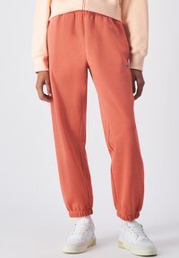 Un pantalon de jogging couleur rouille avec une taille élastique et des poignets, fabriqué en tissu doux. Porté avec des baskets blanches et des chaussettes de cheville.