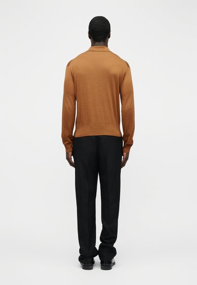 Άνδρας με κοντά σγουρά μαλλιά, που φοράει πορτοκαλί μακρυμάνικο πουκάμισο και μαύρο παντελόνι, στέκεται με την πλάτη προς το μέρος μας σε απλό φόντο.