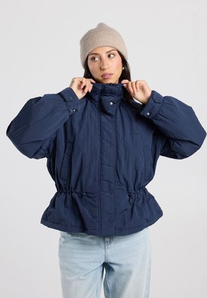Femme portant un bonnet beige en tricot et une doudoune bleu marine avec taille ajustable, remontant le col, debout devant un fond clair uni.