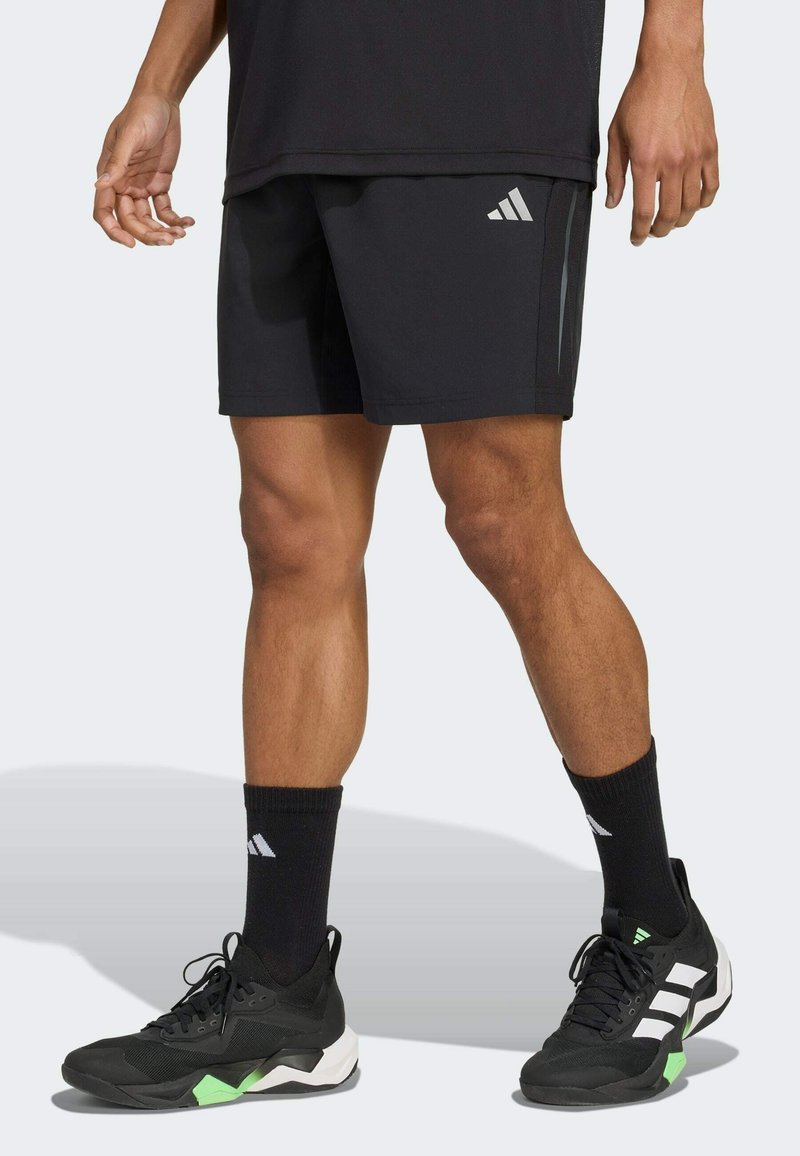 Sorte atletiske shorts med grå Adidas-logo, parret med sorte sokker og sko med grønne accenter og hvide striber.