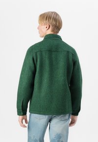 Veste en laine verte avec une surface texturée, col pointu et poignets boutonnés. Présente des fentes latérales et un ourlet droit. Portée avec un jean bleu clair.