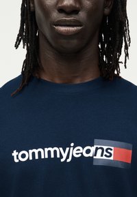 Κοντινό πλάνο ενός ατόμου που φορεί ένα σκούρο μπλε πουκάμισο με το κείμενο «tommyjeans» και ένα κόκκινο, λευκό και μπλε ορθογώνιο λογότυπο στο στήθος.