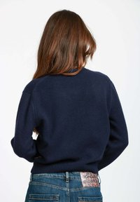 Donna con lunghi capelli castani che indossa un maglione blu navy e jeans blu, con un evidente patch in pelle marrone del marchio Dondup sulla vita.