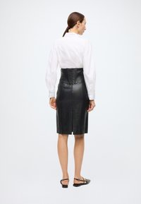 Saia midi de couro preto com silhueta ajustada, apresentando um fecho de correr na parte de trás e acabamento texturizado, combinada com uma camisa branca de mangas compridas.