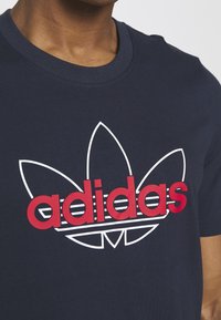 Marinblå bomull t-shirt med en stor röd och vit Adidas trefoil-logotyp, med horisontella ränder under texten. Korta ärmar och rund hals.