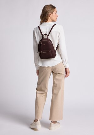 Zaino bordeaux scuro con forma arrotondata, dotato di una tasca frontale con zip, hardware dorato e spalline regolabili. Indossato sopra una camicia bianca.