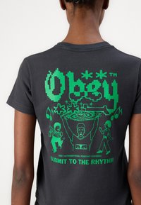 Svart bomulls T-skjorte med lys grønn grafisk trykk av figurer og teksten "Obey" og "Submit to the Rhythm" på baksiden. Korte ermer.