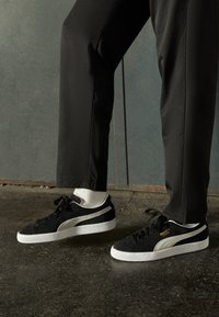Baskets en suede noir avec accents blancs, semelles en caoutchouc texturé et design à lacets, accompagnées de chaussettes de couleur claire et de pantalons noirs.
