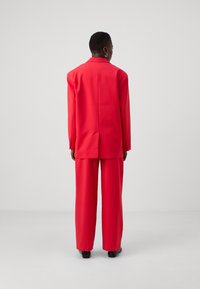 Red tailored suit featuring a long jacket and wide-legged trousers, both in a smooth fabric. The jacket has a single vent and buttoned cuffs.