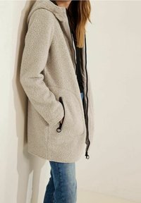 Chaqueta de forro polar gris claro con capucha, con cremallera, dos bolsillos y superficie texturizada. Combinada con vaqueros azules.