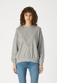 Sweatshirt gris avec des détails en dentelle texturée en forme de V sur le devant, poignets et ceinture côtelés. Accompagné de jeans bleus.