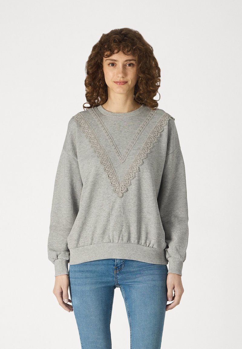 Sweatshirt gris avec des détails en dentelle texturée en forme de V sur le devant, poignets et ceinture côtelés. Accompagné de jeans bleus.