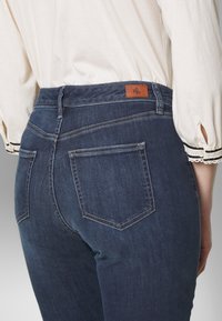 Dark blue denim jeans feature a high waist, two back pockets, and a leather label with initials at the back. The fabric has a smooth texture.