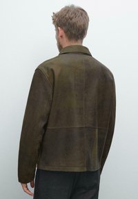 Braune Wildlederjacke mit klassischem Kragen, Rückenpassennaht und langen Ärmeln. Glatte Textur und minimalistisches Design, ohne sichtbare Verschlüsse.