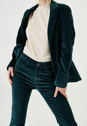 Person iført en teal kordfløjlsdragt med blazer og bukser, kombineret med en struktureret cremefarvet højhalset top.