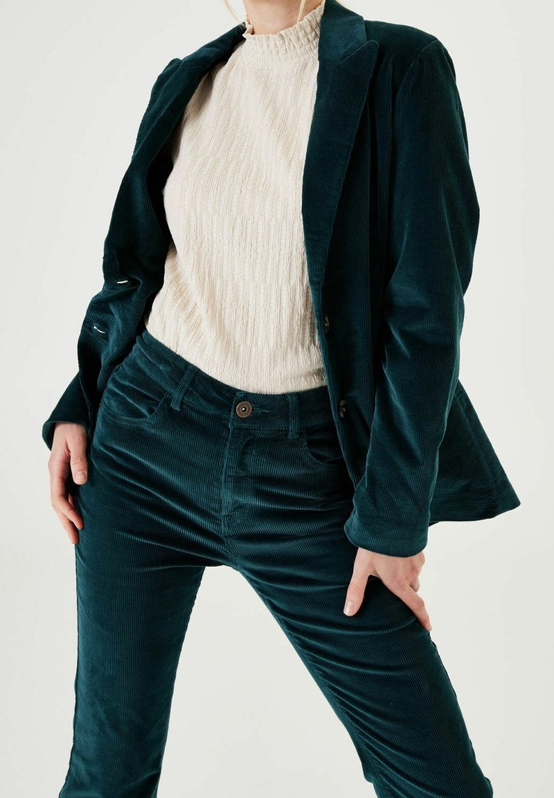 Personne portant un costume en velours côtelé bleu sarcelle avec blazer et pantalon, associé à un haut texturé crème à col montant.