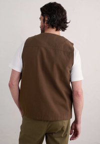 Brązowa bezrękawnik na białej koszulce, o prostym kroju z napinaczem z tyłu i gładką fakturą, w zestawie z oliwkowymi spodniami.