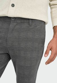 Pull beige rentrée dans un pantalon gris texturé à boutons avec passants pour ceinture, montrant une main droite détendue à côté du pantalon.
