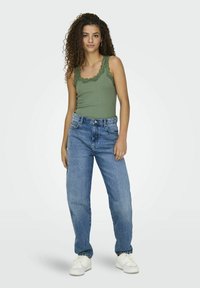 Groene tanktop met kantafwerking, gecombineerd met een ontspannen grijze denim jeans en witte sneakers. Model staat tegen een neutrale achtergrond.