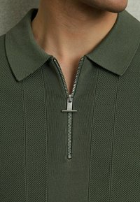 Olive zelena polo majica z teksturiranim tkanjem, ki ima zadrgo in raven ovratnik, poudarja vertikalne črte.
