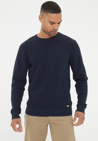 Marineblaue gestrickte Pullover mit Rautenmuster, runder Ausschnitt und gerippten Bündchen. Kleine Markenkennung unten links.