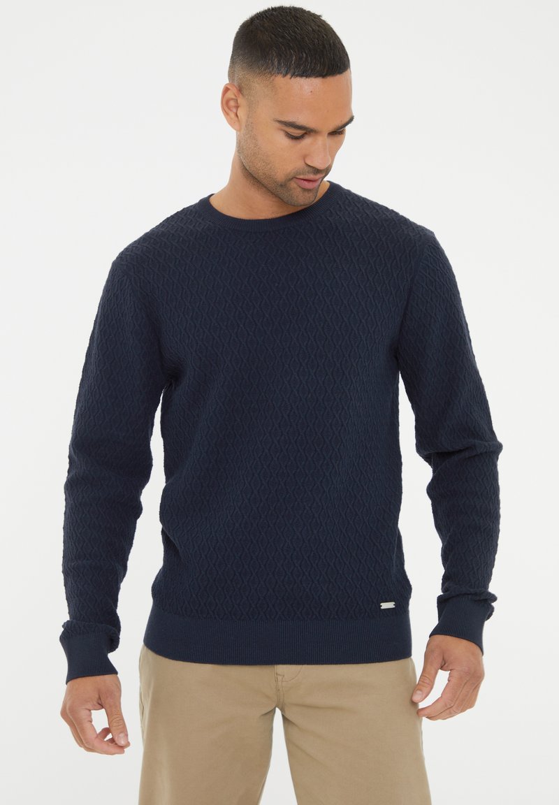 Marineblaue gestrickte Pullover mit Rautenmuster, runder Ausschnitt und gerippten Bündchen. Kleine Markenkennung unten links.