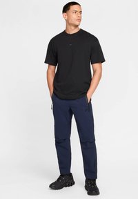 Czarny t-shirt z krótkim rękawem z małym czarnym logo na piersi, zestawiony z granatowymi spodniami typu cargo z kieszeniami na zamek oraz czarnymi sportowymi butami.