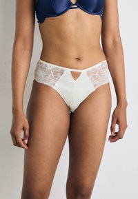 Höga beige underkläder med spetspaneler på sidorna och en triangelutskärning framtill, med en slät satinliknande textur.